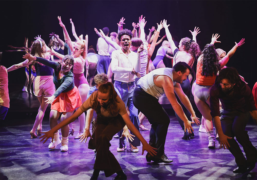 Full ensemble cast dances energetically under purple stage lighting in 1970s rehearsal wear, A Chorus Line, National Theatre St Kilda.