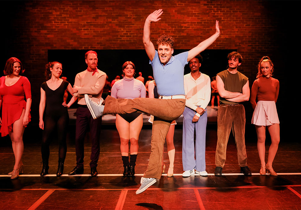 A performer in a blue polo kicks high mid-dance move while lined up alongside castmates against a brick backdrop.