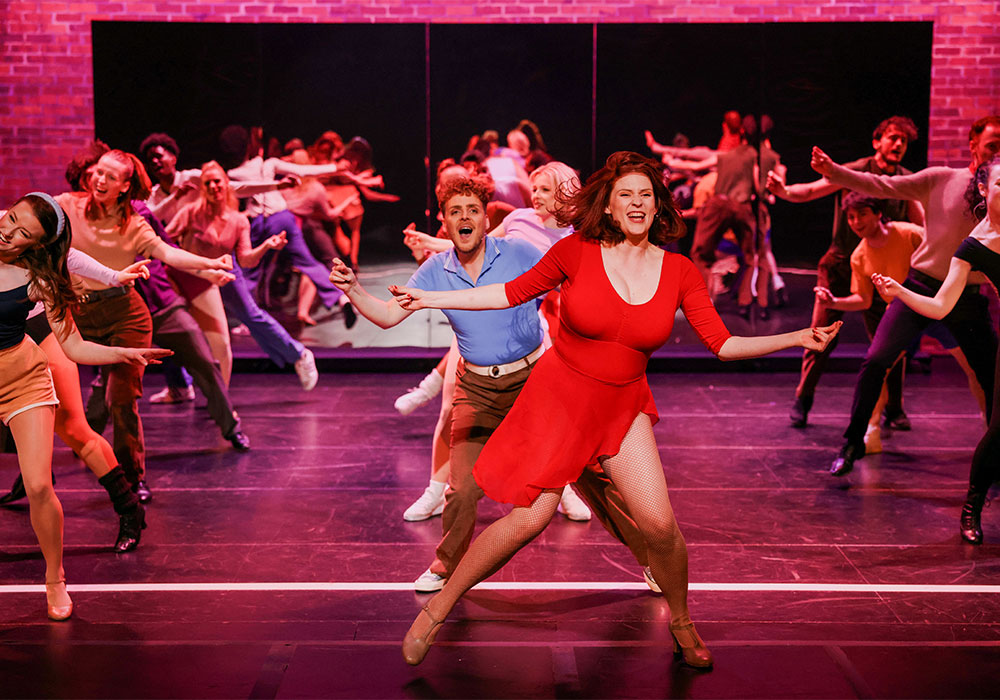 A performer in a red dress dances passionately centre stage as the ensemble moves behind her, opened 7 September 2024.