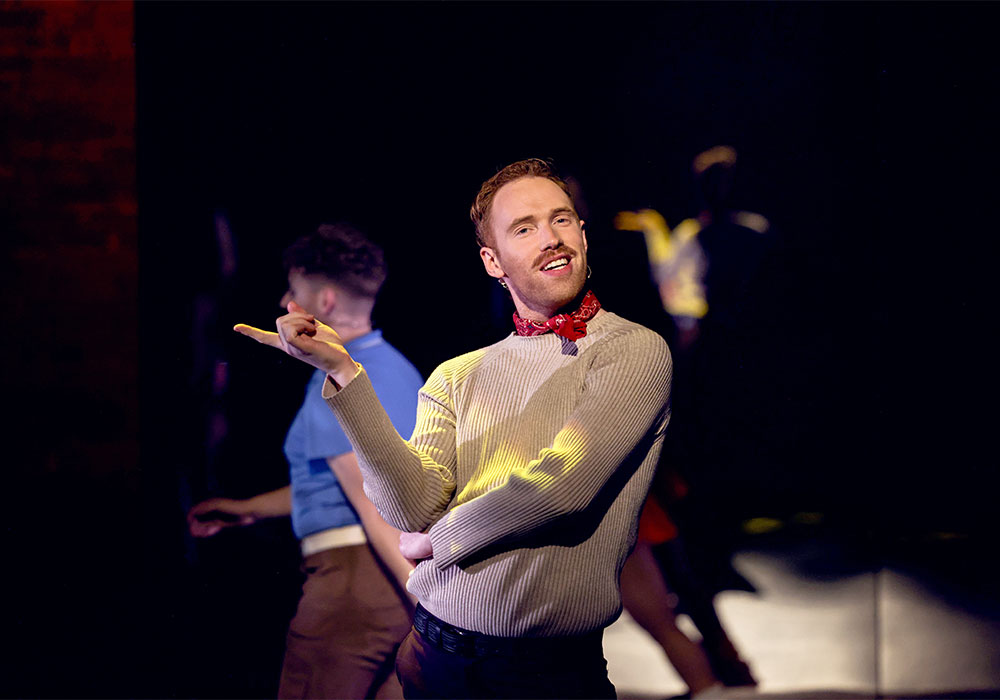 A red-headed performer in a beige jumper and red bandana strikes a playful, confident pose mid-scene.