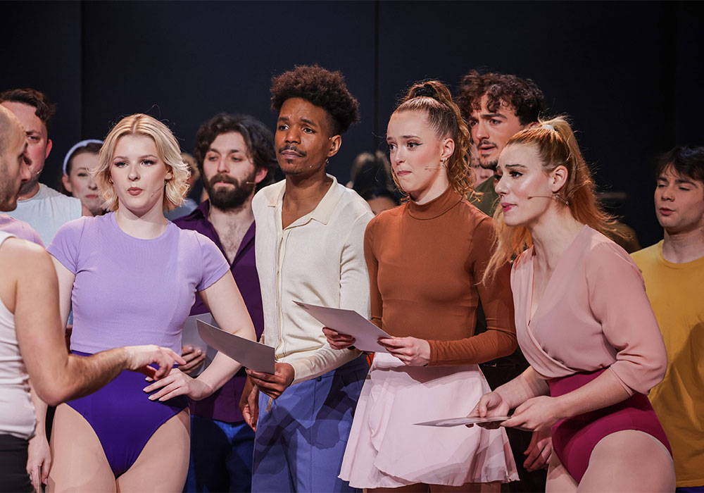 Cast members in colourful rehearsal outfits stand together holding audition sheets, portraying nervous dancers awaiting their fate.