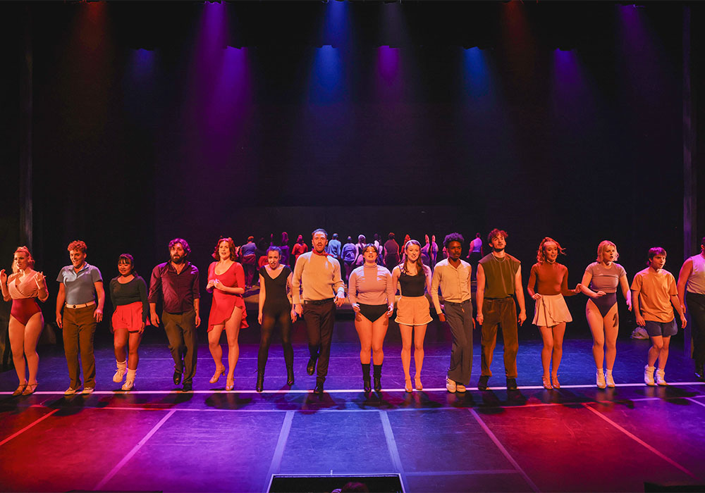 The full company lines up together on stage under red and blue lighting for a striking finale pose.