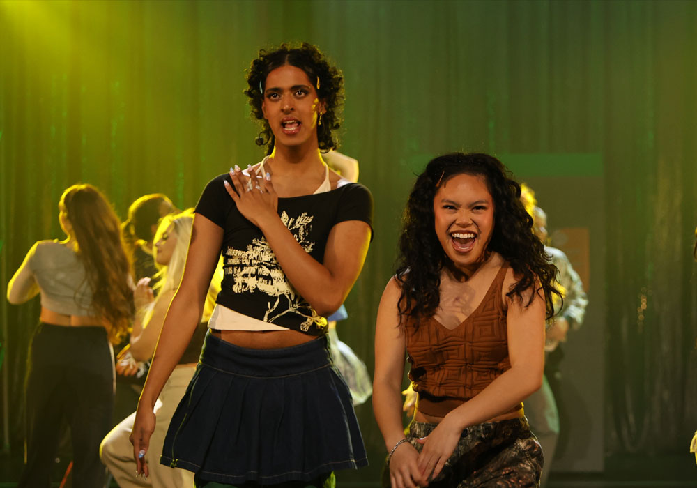 Two performers dance energetically centre stage, one in a black graphic tee and denim mini skirt, the other laughing in a brown top, with ensemble dancers blurred behind them.
