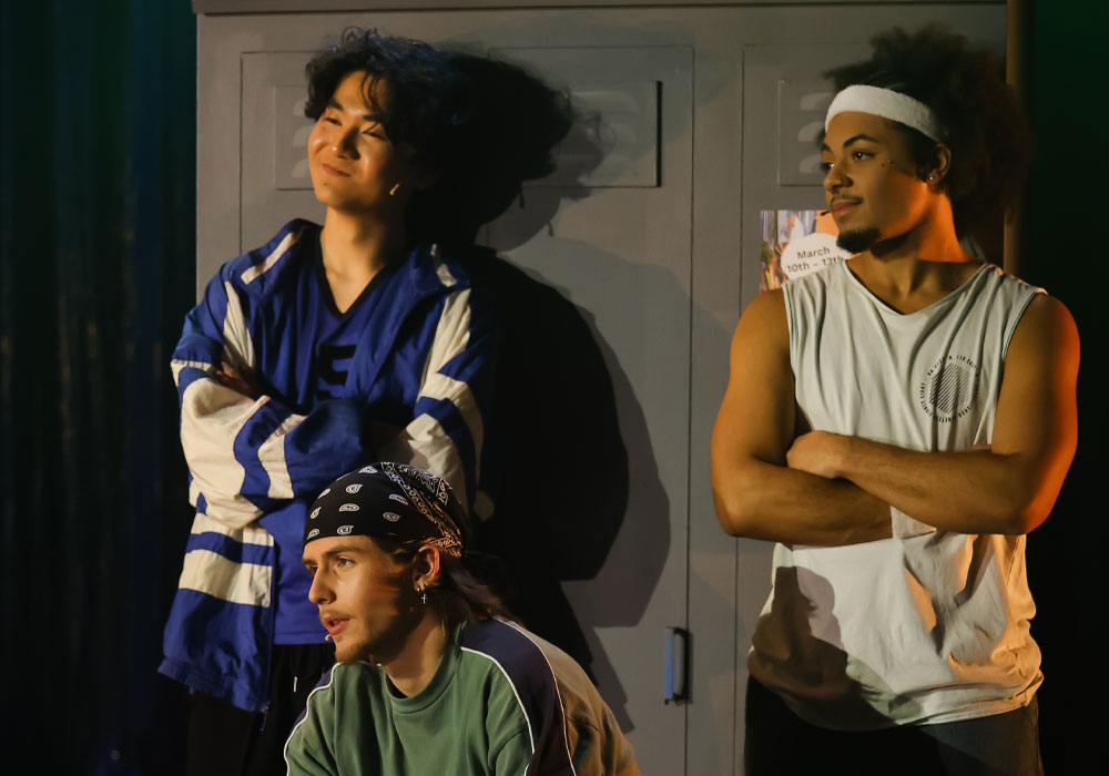 Three male cast members in a locker room scene from Bring It On at the National Theatre St Kilda: one leans against the lockers in a varsity jacket, one kneels wearing a bandana, and one stands with arms crossed in a headband.