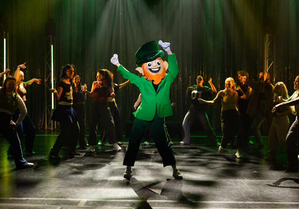 A comic leprechaun mascot dances centre stage with arms raised, surrounded by the energetic Bring It On ensemble cast.