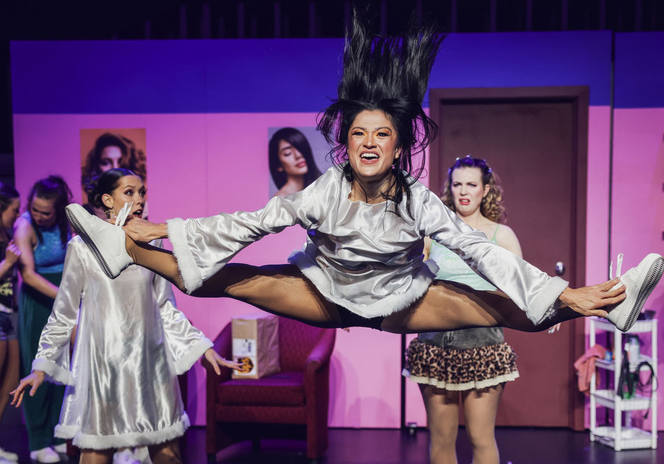 A performer in a white satin robe leaps mid-air doing the splits during a comedic salon scene, surrounded by fellow cast members.