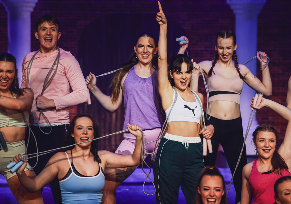 Ensemble cast in gym gear skip rope and cheer together during an energetic workout number, an ANDREW G and Theatrical production.