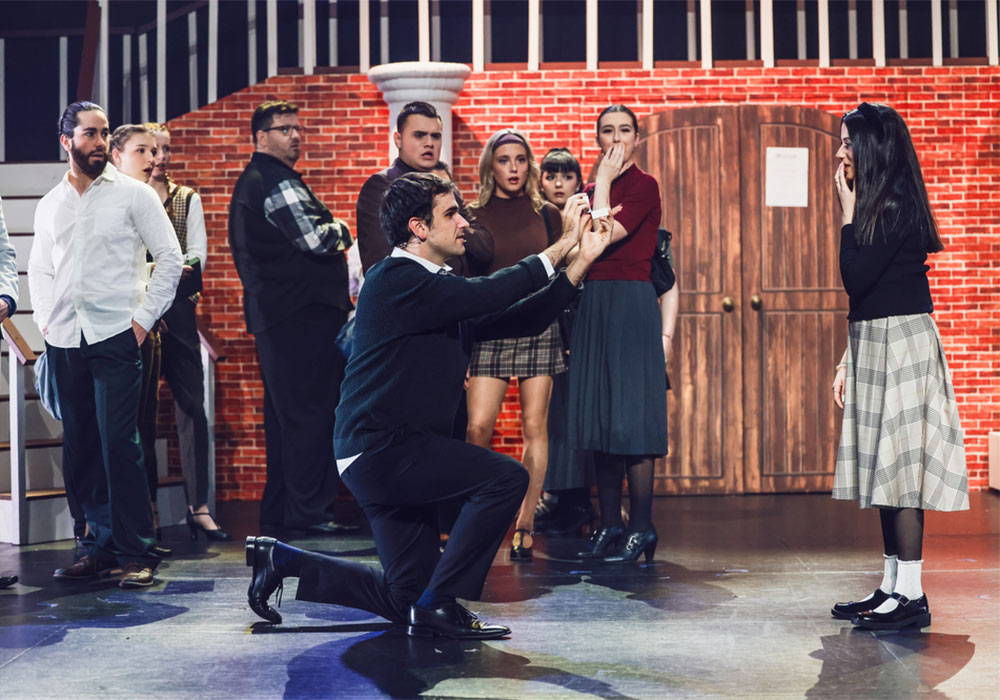 A man kneels to propose in front of a stunned ensemble cast dressed in preppy 90s attire, licensed exclusively by Music Theatre International (Australasia).