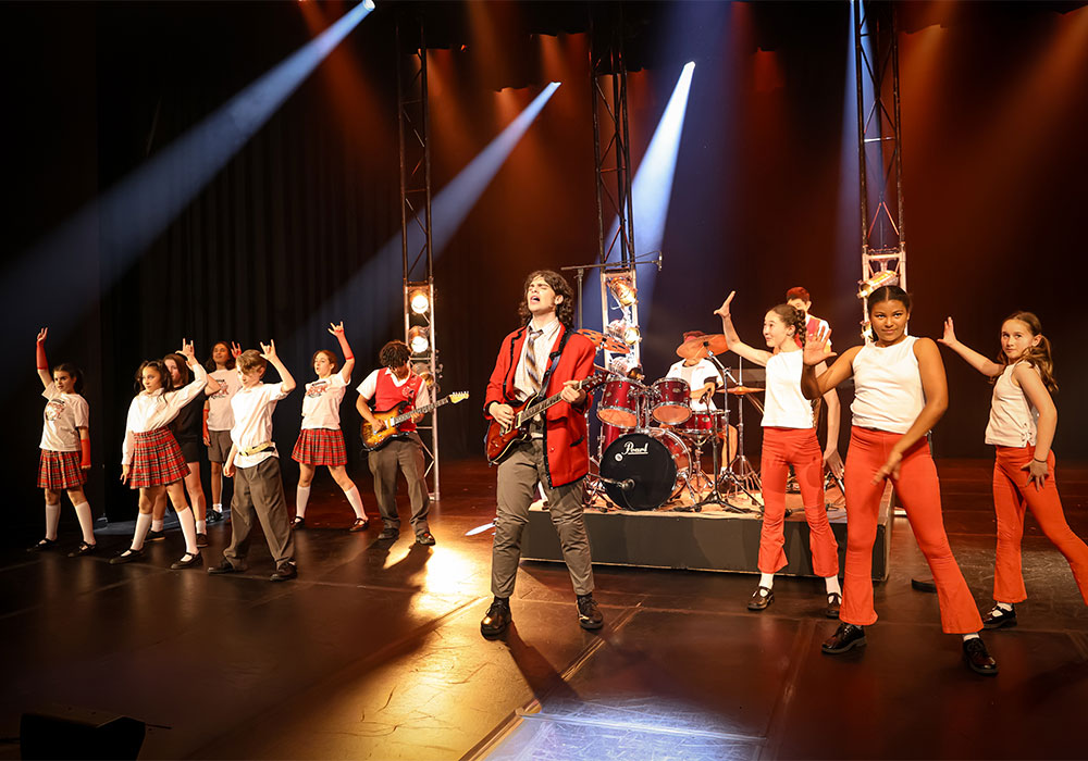 Student rock band performs on stage with electric guitars and drums as classmates in school uniforms cheer, School of Rock musical, National Theatre St Kilda, November 2025.