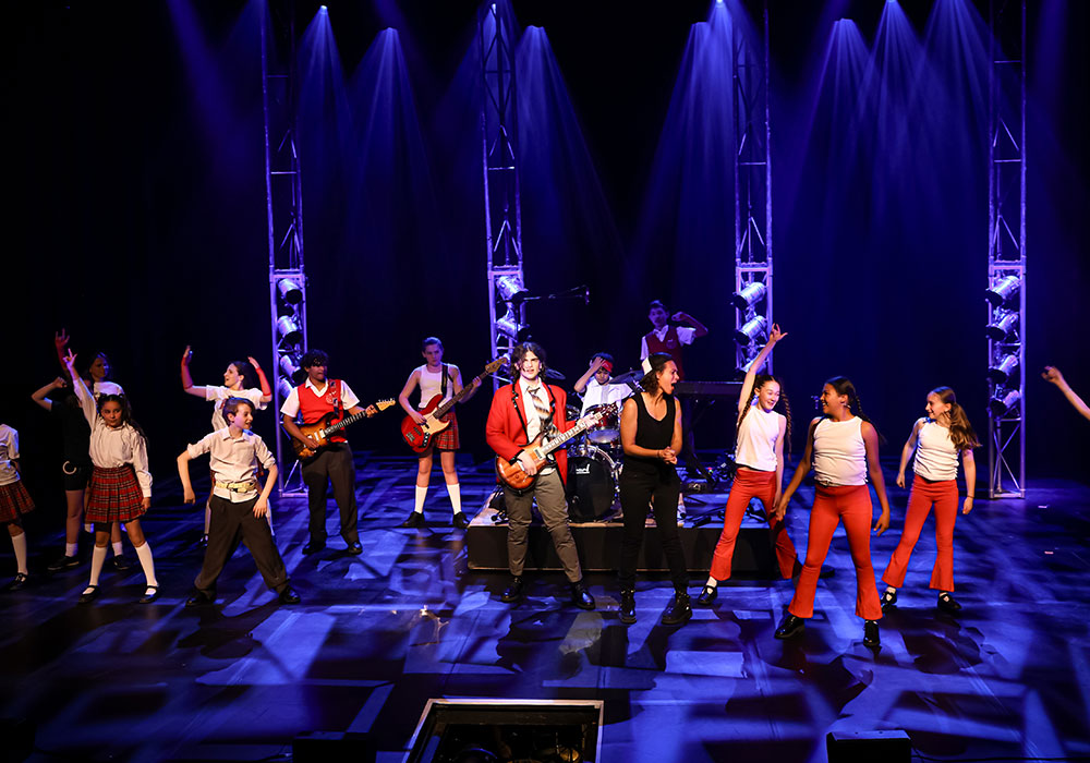 The full cast of School of Rock dances under blue stage lighting during a high-energy band performance, an ANDREW G and Theatrical production.
