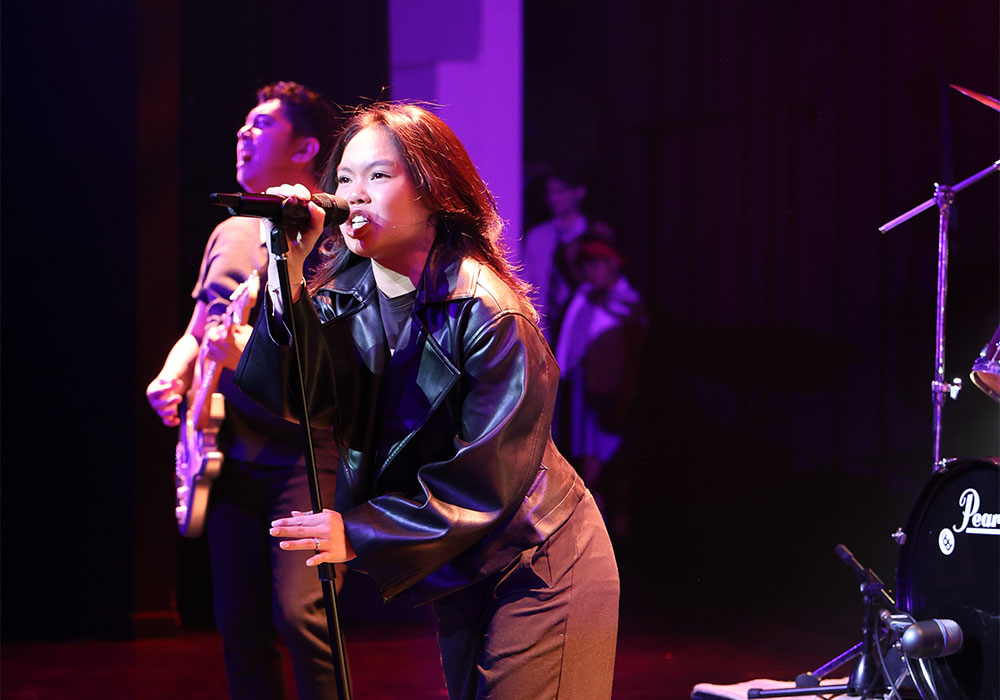 A young performer sings passionately into a handheld microphone beside an electric guitarist, lit in purple stage lighting.