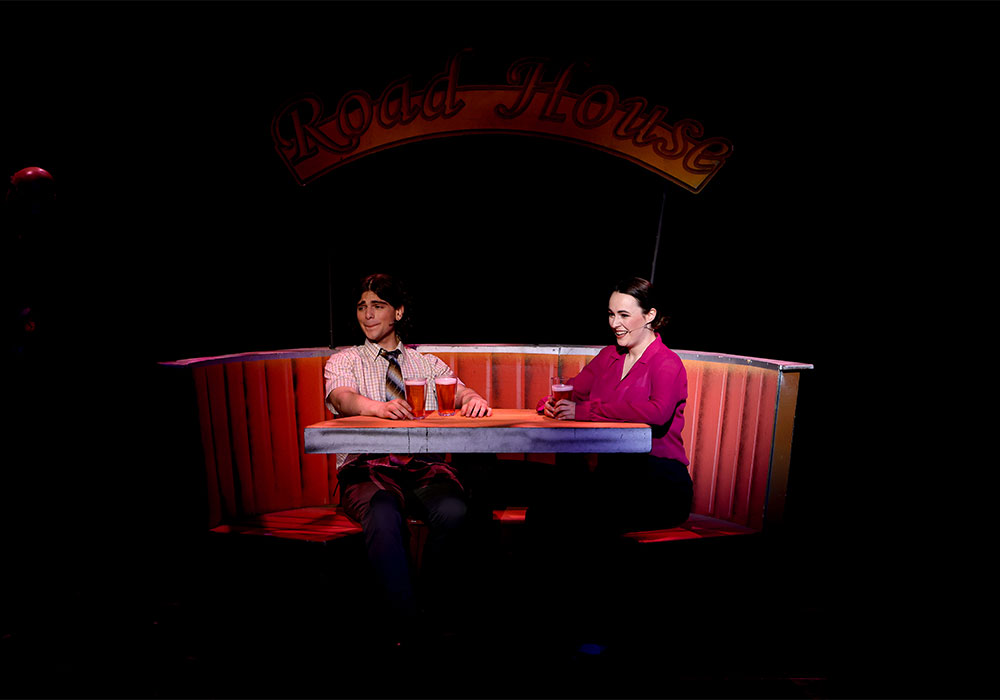 Two actors sit across from each other at a "Road House" diner booth, sharing drinks in a warmly lit scene from School of Rock.